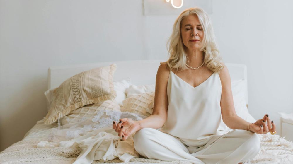Woman in White Strap Top Sitting on Bed in Lotus Pose with Eyes Closed | Peak Performance Aging